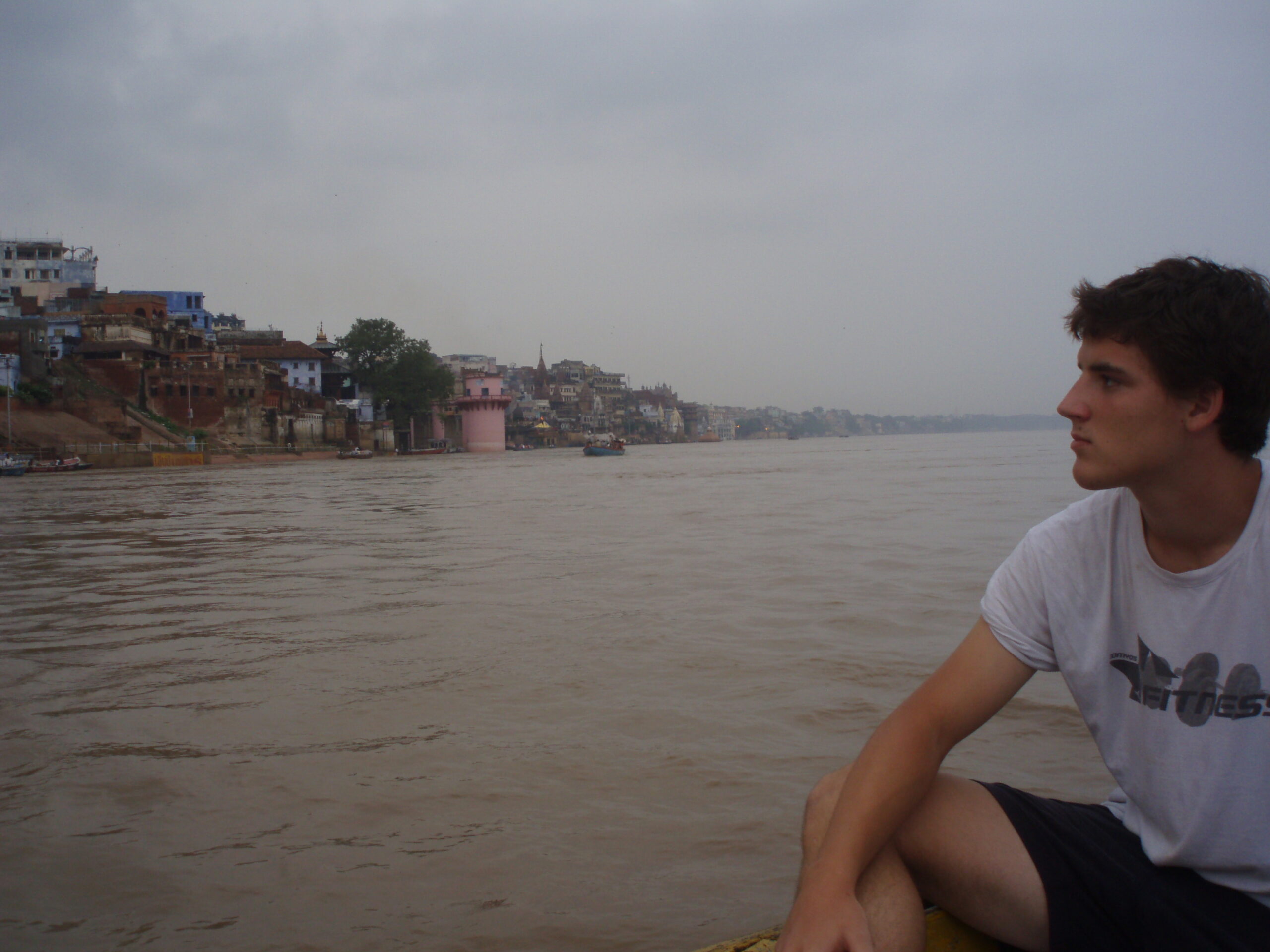 Un joven sentado en una barca contempla el río, con los gates y edificios de una ciudad india al fondo