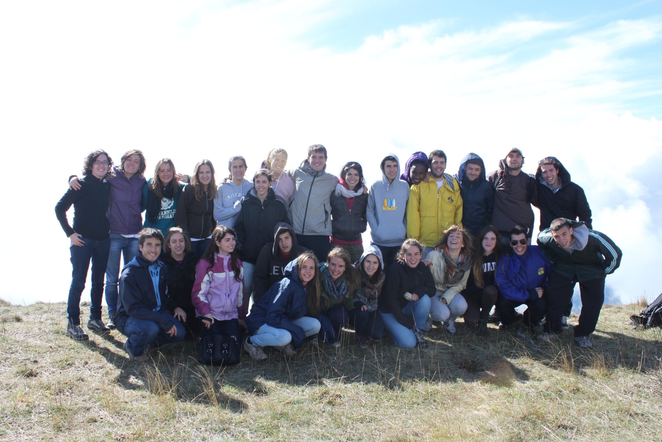 Um grupo numeroso de amigos com Marcos no alto de uma montanha, sobre um mar de nuvens