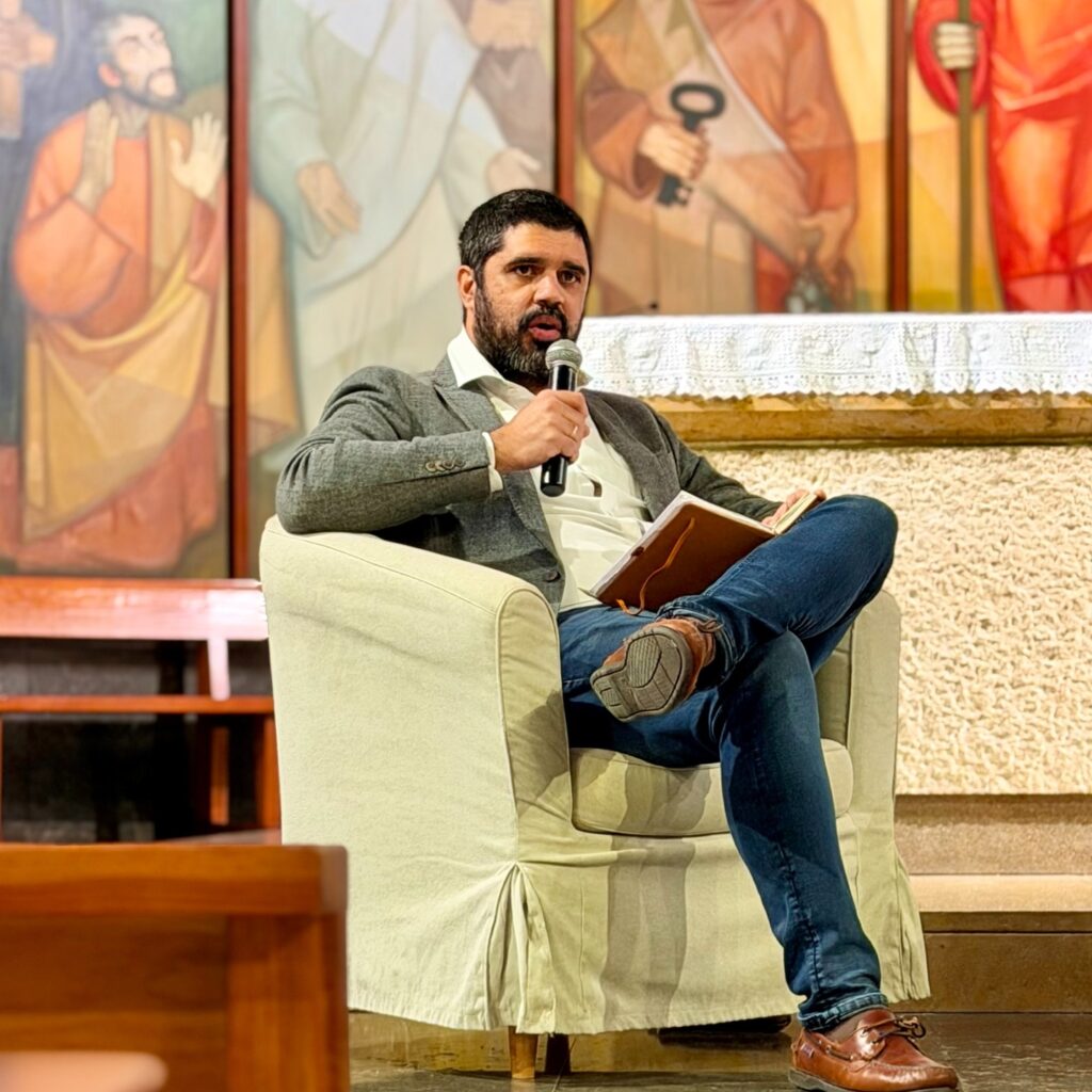 Alfonso Calavia speaks into a microphone, seated in an armchair with an open book on his lap