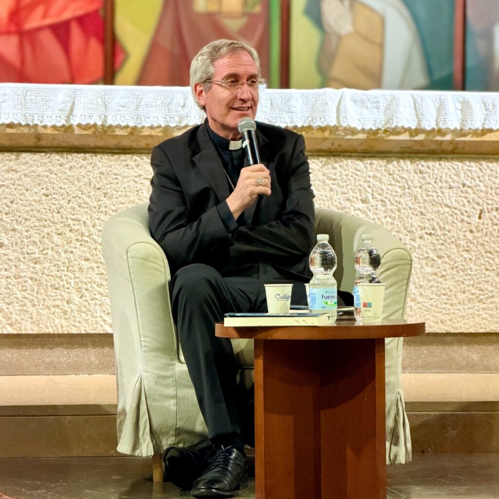 Mons. Javier Vilanova speaks into a microphone, seated beside a low table with glasses and bottles of water