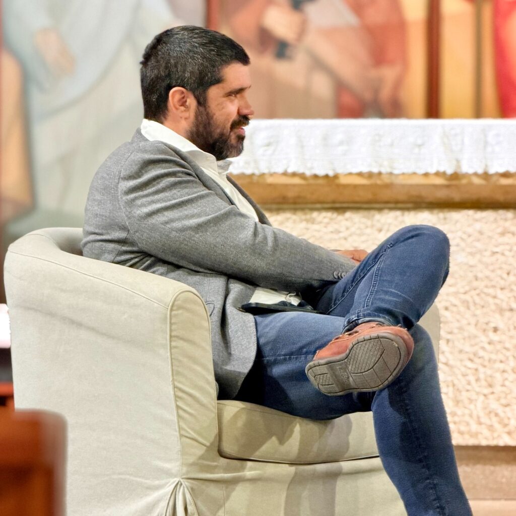 The author listens seated in the armchair, in profile, before the altar