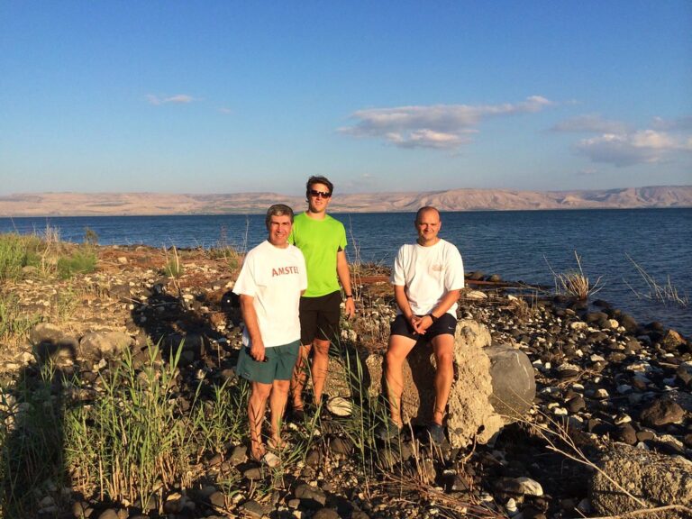 Marcos Pou con dos hombres en la orilla pedregosa de un lago, con montañas al fondo y cielo azul