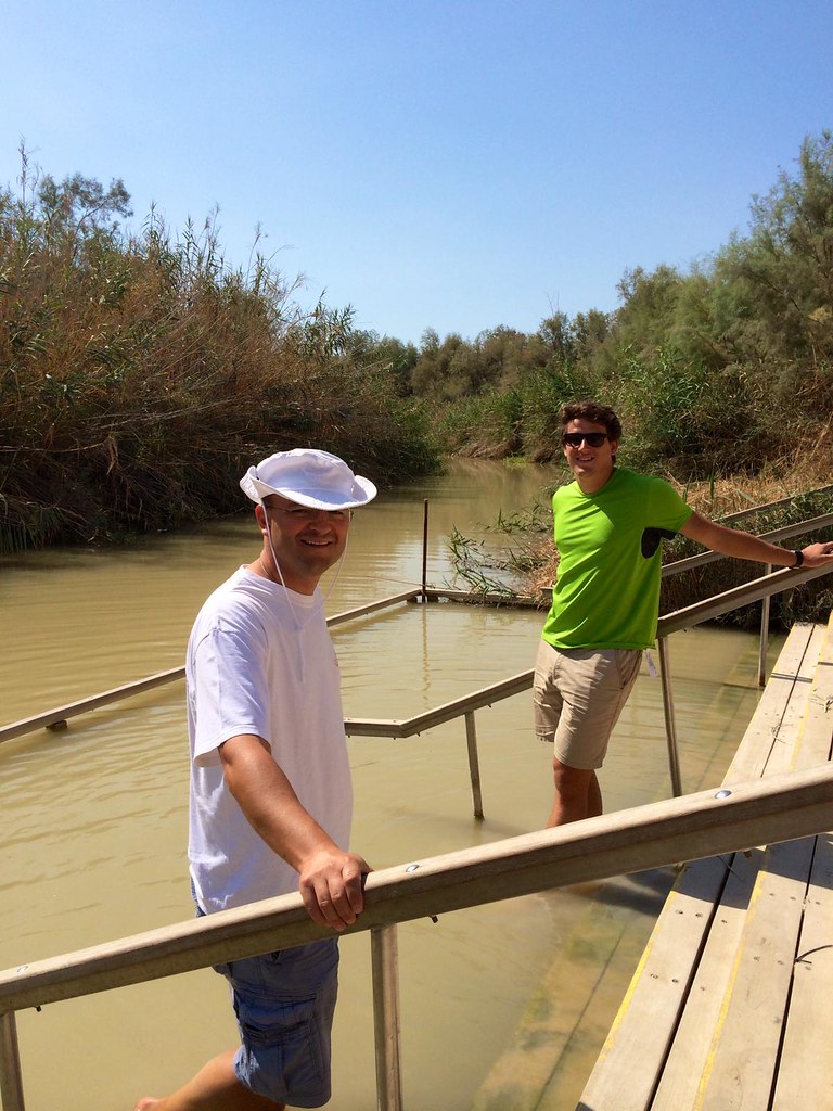 Dos jóvenes sonrientes bajan por una pasarela de madera hacia las aguas del río Jordán en un día soleado