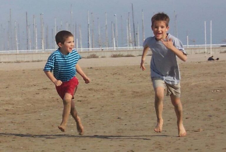 Dos niños descalzos corriendo y sonriendo por la arena de una playa, con mástiles de barcos al fondo