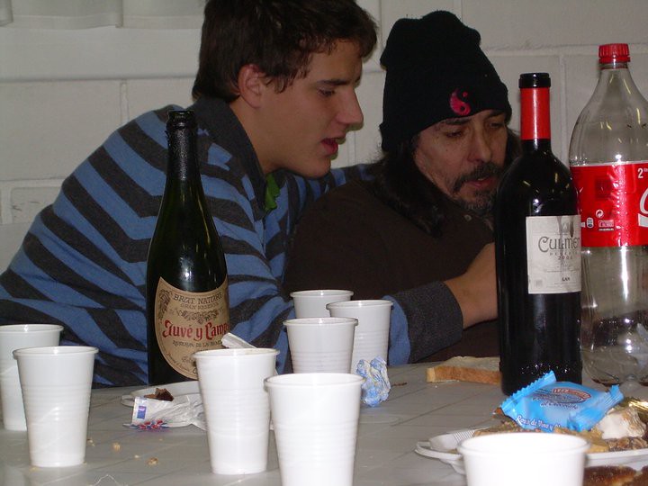 Marcos Pou conversando con un hombre con gorro en una mesa con vasos, botellas y comida durante una comida solidaria