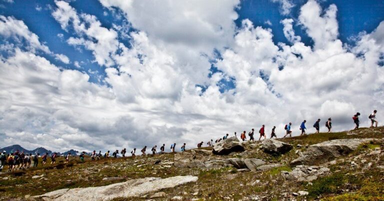 Larga fila de excursionistas con mochilas caminando por una cresta rocosa de montaña bajo un cielo de grandes nubes