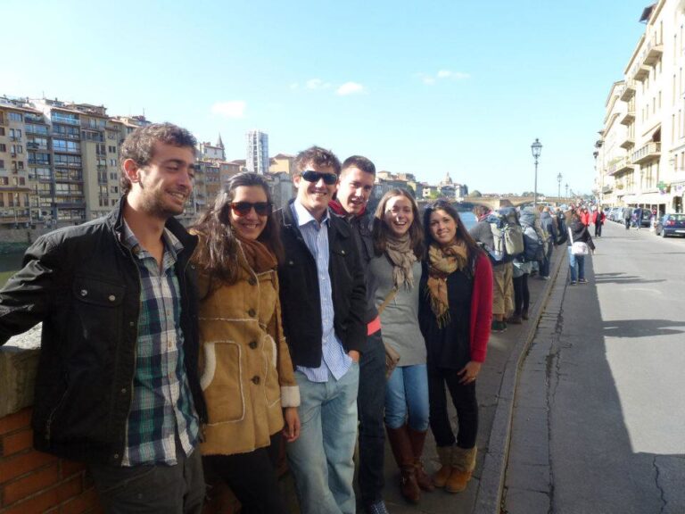 Grupo de jóvenes sonrientes alineados junto al pretil de un río en una ciudad italiana, con el Ponte Vecchio al fondo
