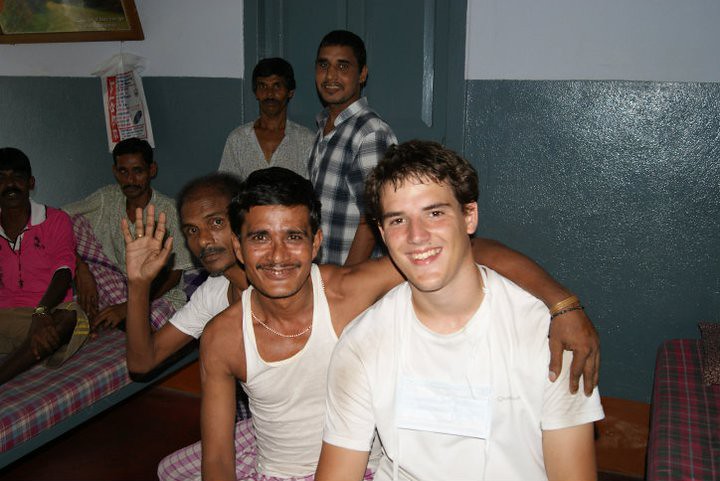 Marcos Pou sonriente y abrazado a un hombre en una sala con varios hombres sentados en camas durante un viaje a la India