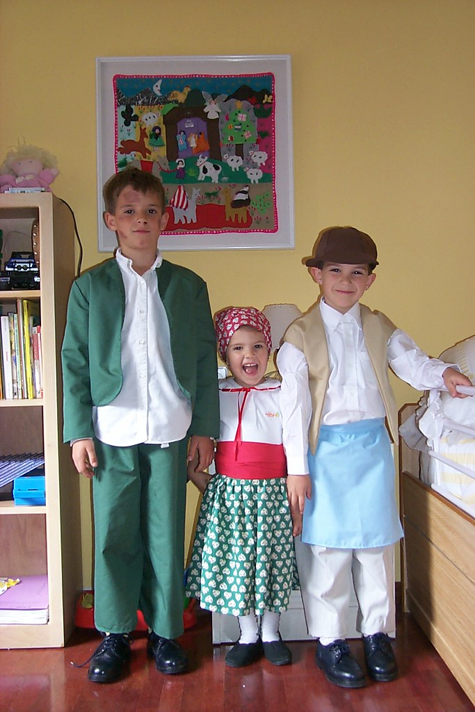 Tres niños disfrazados con trajes tradicionales posan en una habitación infantil junto a una estantería con libros
