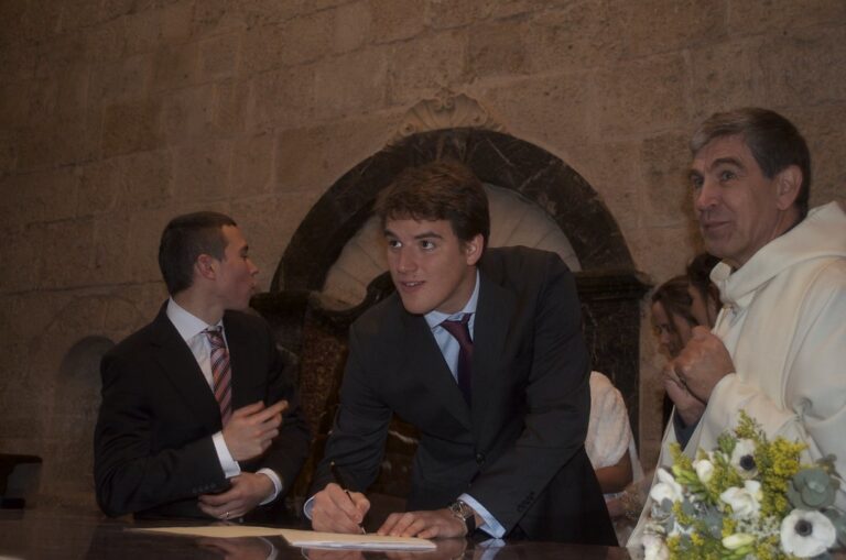 Marcos Pou, de traje, firma un documento en una iglesia junto a un joven, un sacerdote y un ramo de flores