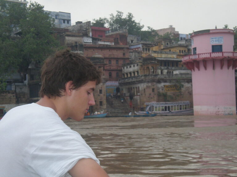 Marcos Pou de perfil, con camiseta blanca, en una barca ante los gaths y edificios a orillas de un río en la India