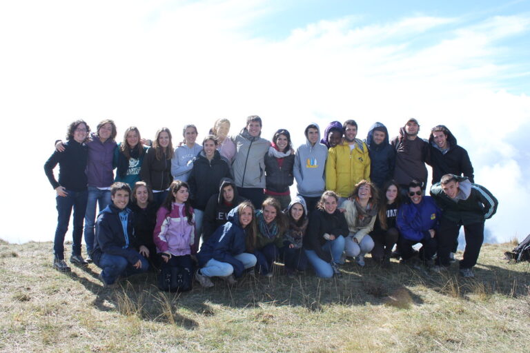 Grupo numeroso de jóvenes posando abrazados en la cima de una montaña, sobre la hierba y por encima de las nubes