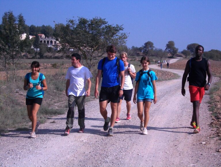 Grupo de jóvenes caminando por un camino de tierra entre campos en un día soleado, con mochilas y ropa deportiva