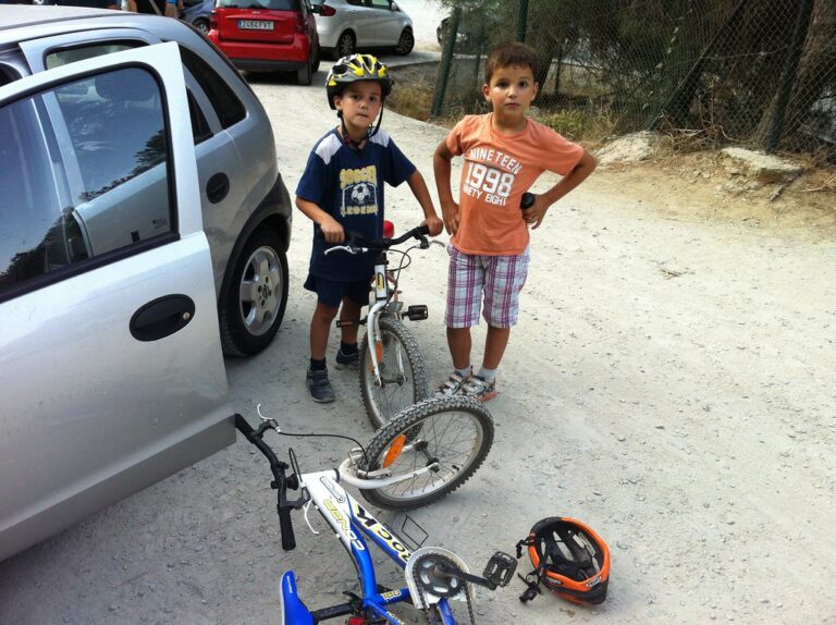 Dos niños con bicicletas junto a un coche en un camino de tierra, uno con casco amarillo y otro con casco naranja en el suelo