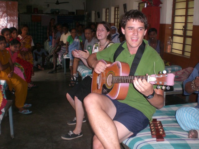 Marcos Pou playing the guitar and singing while seated on a bed, surrounded by residents and volunteers in a mission house in India