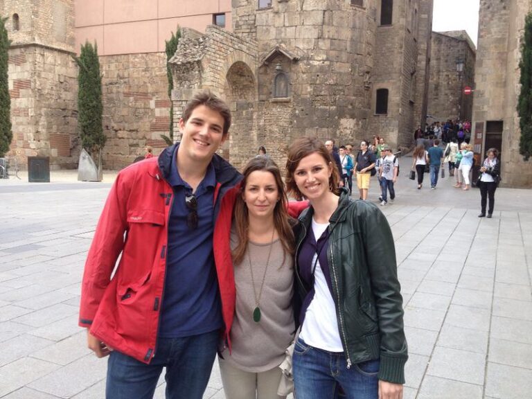 Marcos Pou con chaqueta roja abrazado a dos jóvenes en una plaza del casco antiguo de Barcelona