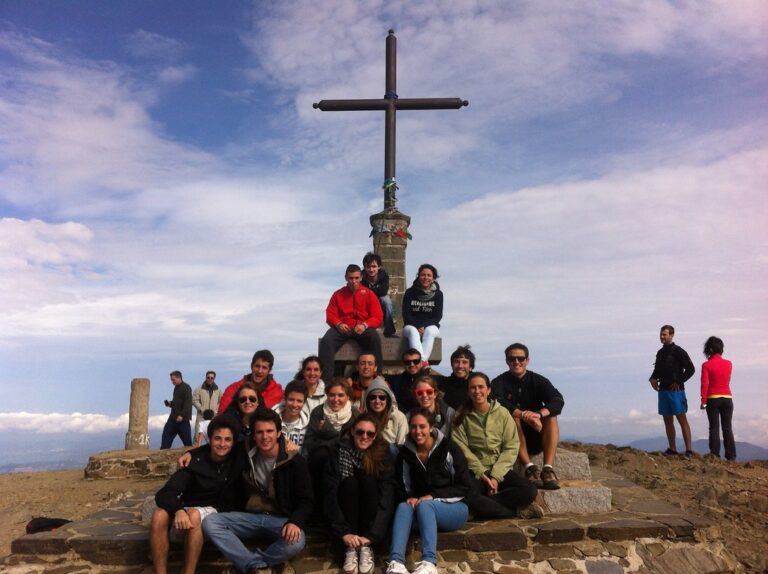 Grupo de jóvenes posando junto a la gran cruz de piedra en la cima de una montaña, con el cielo nublado de fondo