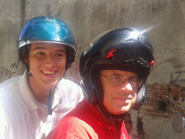 Marcos Pou sonriente con casco azul sobre una moto, junto a un hombre con casco negro y camiseta roja, ante un muro de piedra