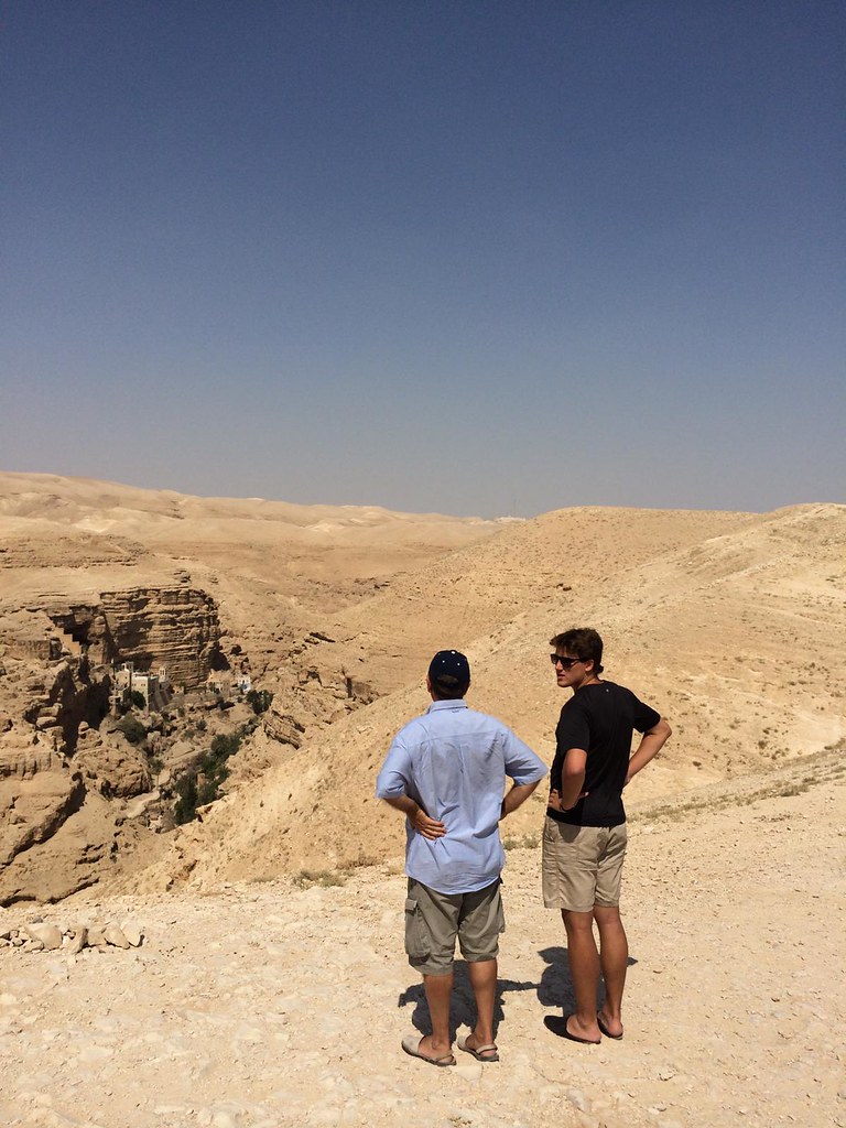 Dos jóvenes de espaldas contemplan un desierto con un monasterio excavado en el acantilado al fondo