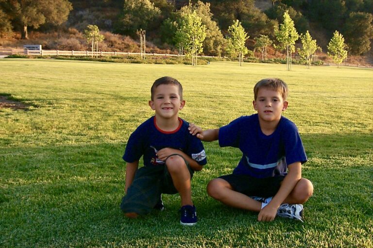 Dos niños con camisetas azules posando sentados sobre el césped de un parque, con árboles y una ladera al fondo