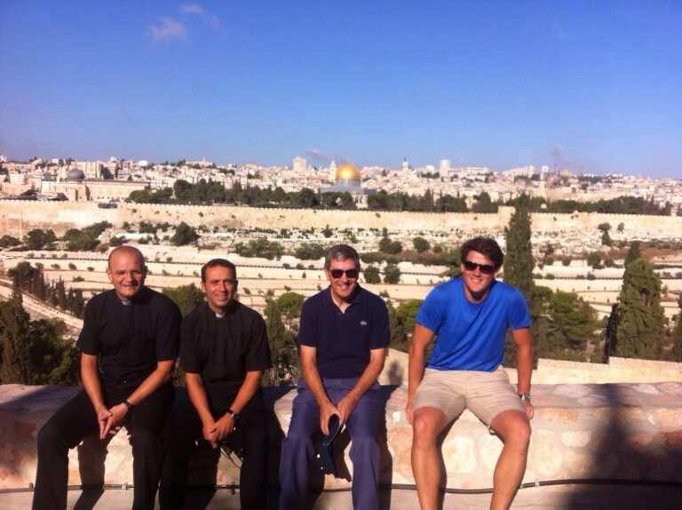 Marcos Pou con dos sacerdotes y otro hombre, sentados en un muro con Jerusalén y la Cúpula de la Roca al fondo