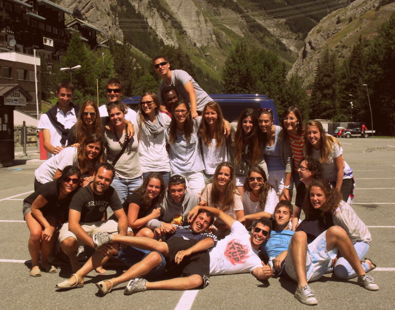 Grupo numeroso de jóvenes posando sonrientes en un aparcamiento de un pueblo de montaña durante un viaje de verano