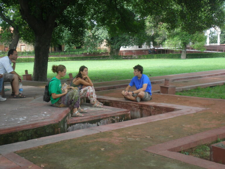 Marcos Pou, sentado en el suelo de un jardín, conversa con dos jóvenes sentadas en un murete bajo los árboles