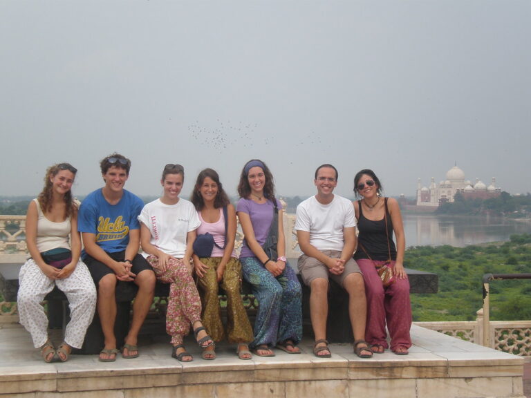 Grupo de jóvenes sentados en un muro sonriendo, con el Taj Mahal y el río al fondo durante un viaje a la India