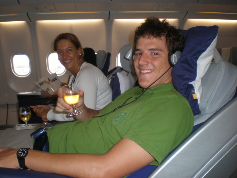 Marcos Pou sonríe en el asiento de un avión con auriculares y una copa en la mano, junto a una mujer que también sonríe