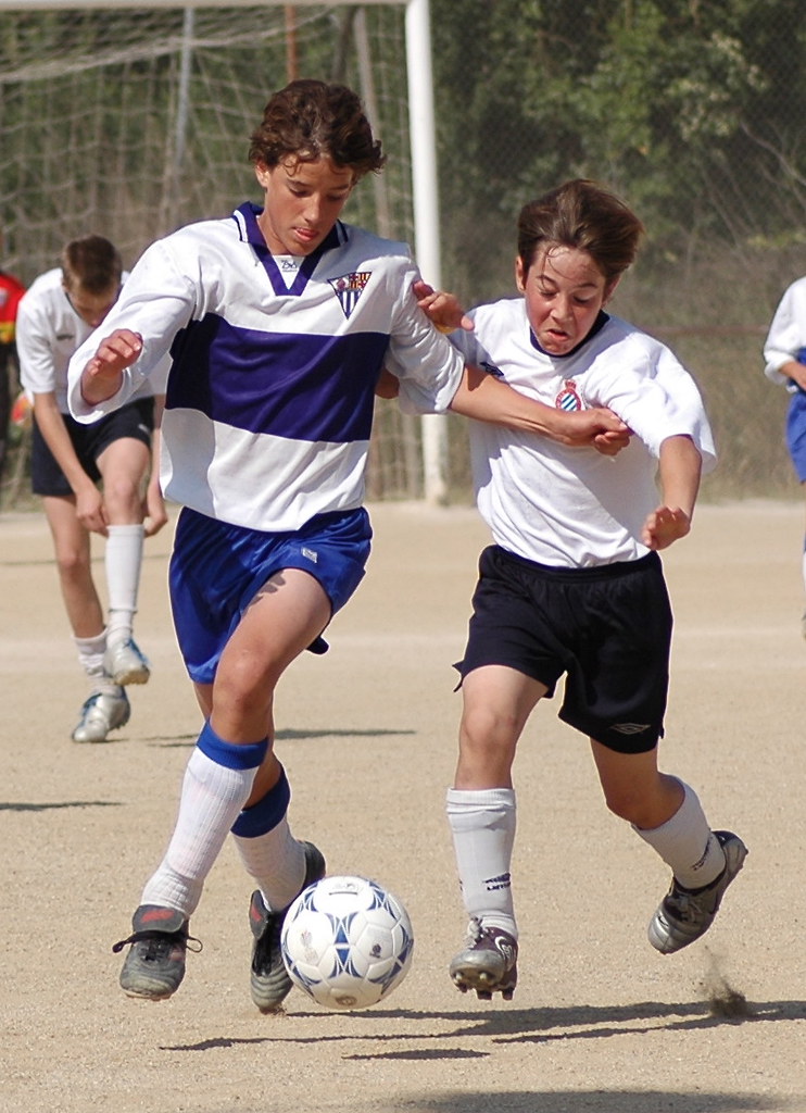Marcos Pou, con camiseta blanca y azul, pugna por el balón con otro jugador durante un partido de fútbol en un campo de tierra