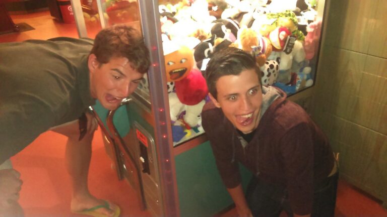 Dos jóvenes hacen muecas divertidas agachados junto a una máquina de peluches en un salón recreativo