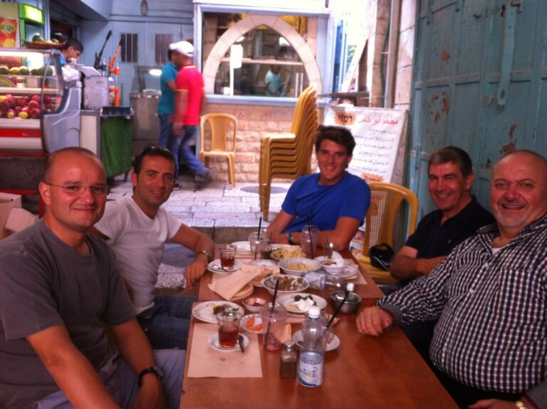 Marcos Pou y un grupo de hombres sonríen sentados a la mesa de un restaurante callejero con platos y bebidas