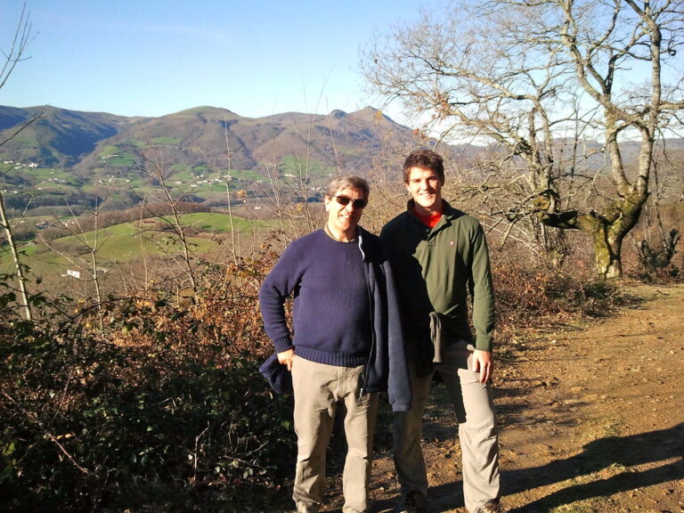Marcos Pou y un hombre mayor posan sonrientes en un camino de montaña, con valles verdes y cumbres al fondo