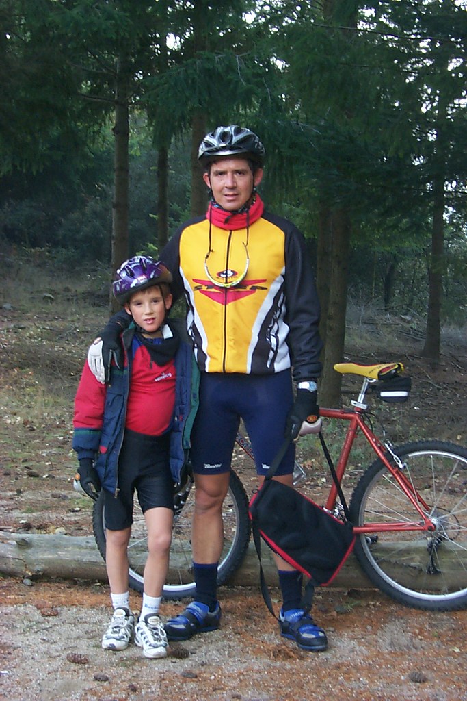 Marcos Pou de niño, con casco y ropa de ciclismo, junto a un hombre adulto y una bicicleta en un bosque de pinos