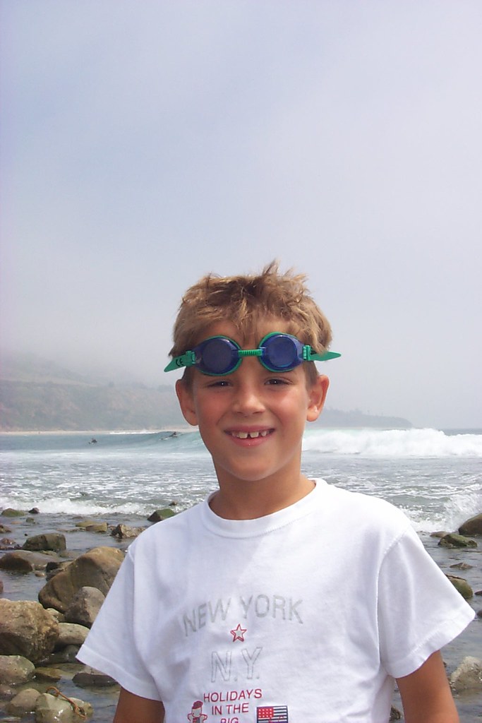 Marcos Pou de niño, sonriente con gafas de bucear en la frente y camiseta blanca, junto al mar y unas rocas