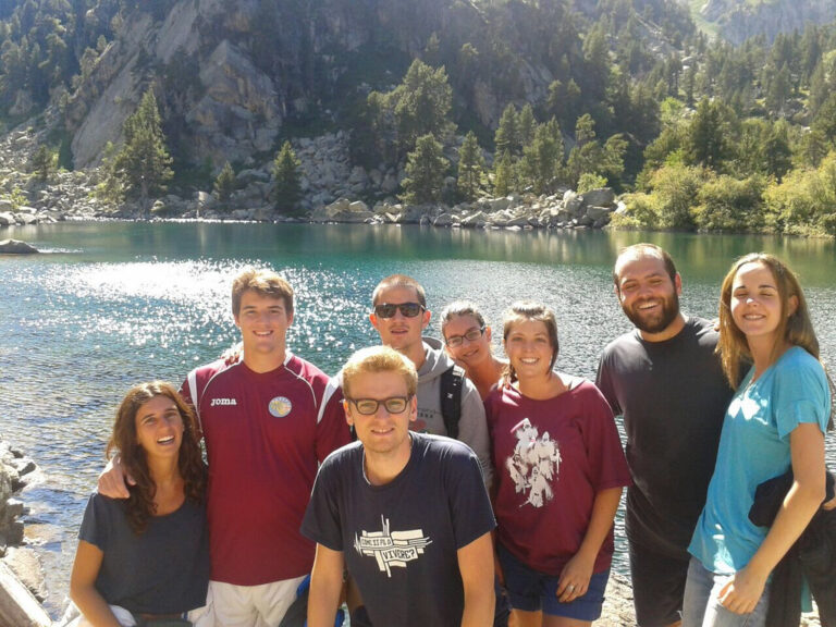 Grupo de ocho jóvenes sonrientes posando juntos junto a un lago de montaña rodeado de pinos y rocas