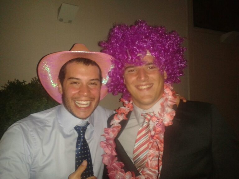 Dos jóvenes sonrientes en una fiesta, uno con sombrero rosa y otro con peluca morada y collar de flores