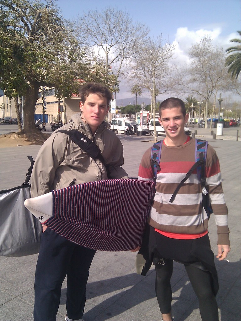Dos jóvenes con tablas de surf y neopreno posando en un paseo junto al mar en un día soleado