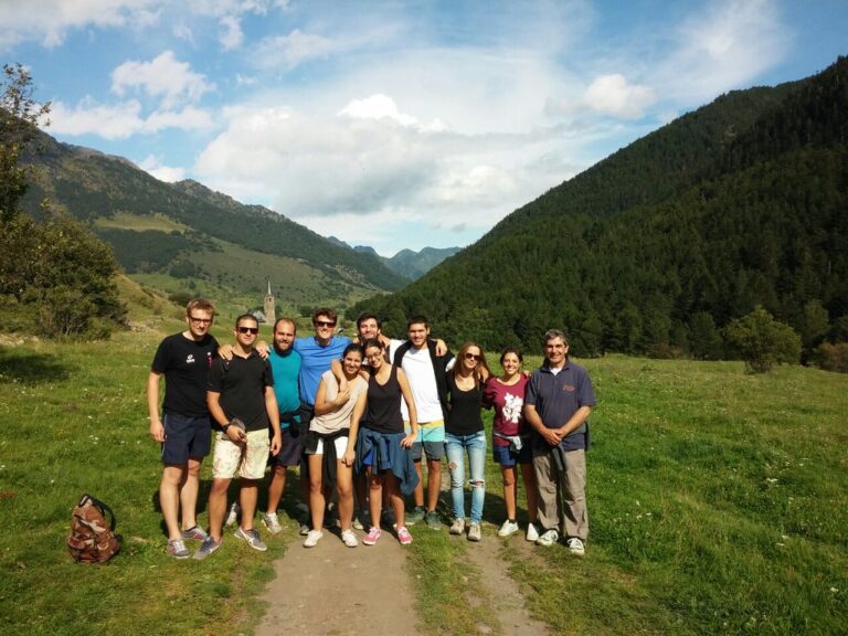Grupo de jóvenes posando en un prado de montaña durante una excursión, con valle boscoso y una ermita al fondo