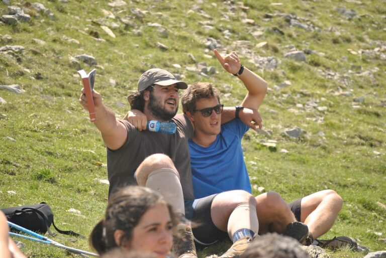 Dos jóvenes sentados en un prado de montaña, uno con gorra y un libro en la mano señalando a lo lejos