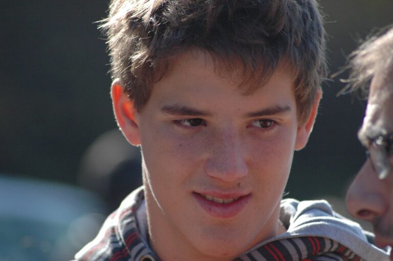 Primer plano de Marcos Pou sonriendo levemente al aire libre, con sudadera de capucha y otra persona a su lado
