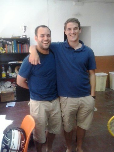 Marcos Pou abrazado por un joven con polo azul, ambos sonrientes en una sala con estanterías de libros