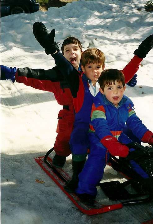 Tres niños con monos de esquí sonríen y levantan los brazos subidos a un trineo sobre la nieve