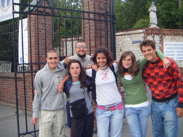 Grupo de seis jóvenes abrazados y sonrientes ante la verja de entrada de un recinto de ladrillo durante un viaje