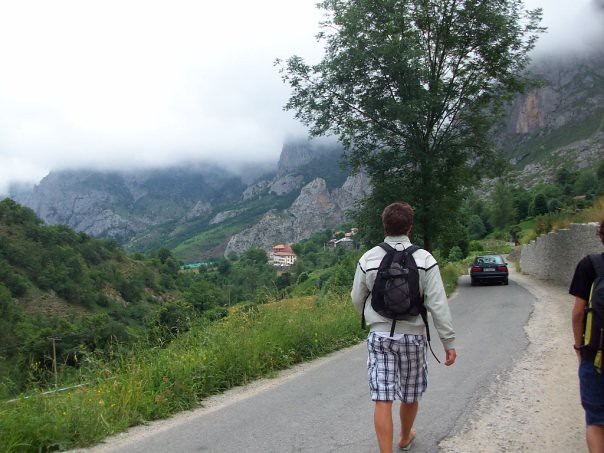 Un joven con mochila camina por una carretera de montaña entre valles verdes y cumbres cubiertas de niebla