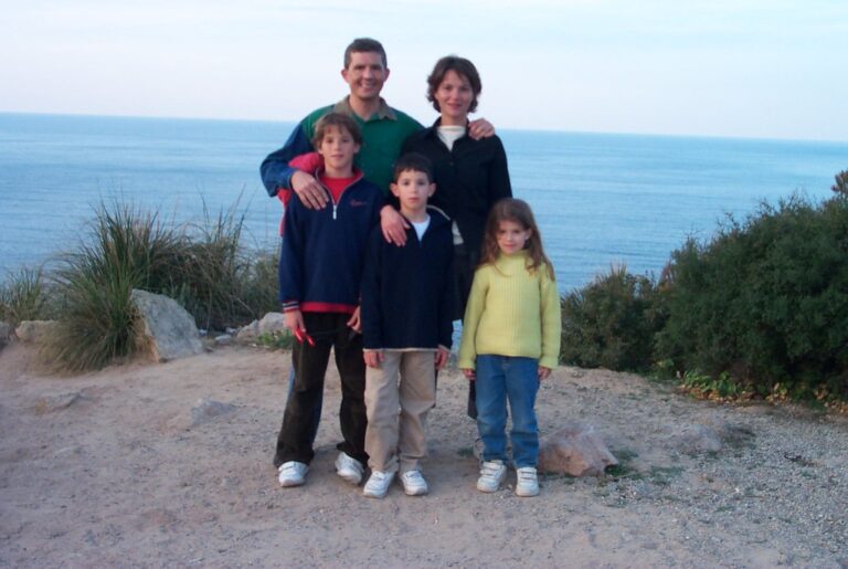 Un matrimonio y tres niños posando juntos en un mirador de tierra junto al mar, con arbustos alrededor