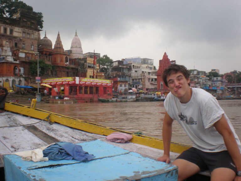 Marcos Pou sentado en una barca haciendo una mueca divertida, con los templos y ghats de un río de la India al fondo