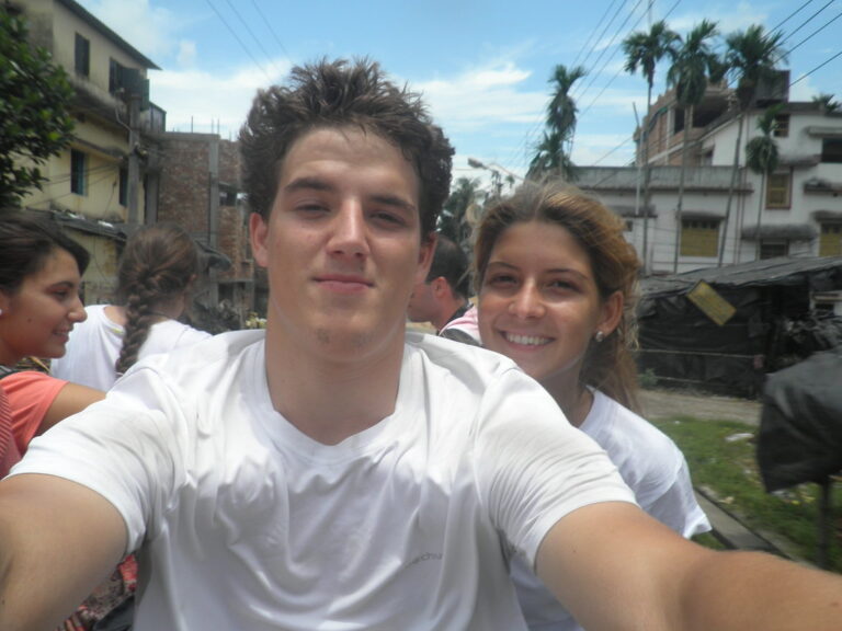Marcos Pou se hace una foto con un grupo de jóvenes en una calle de un país tropical, con palmeras y casas al fondo