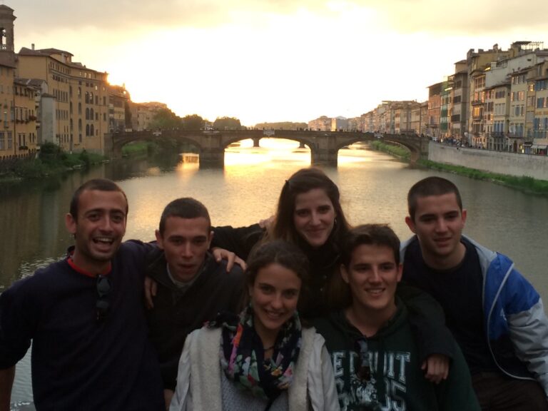 Grupo de seis jóvenes sonrientes abrazados sobre un puente del río Arno en Florencia al atardecer
