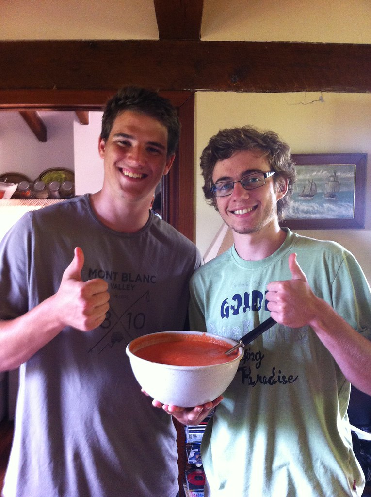 Dos jóvenes sonrientes levantan el pulgar mientras sostienen un cuenco de gazpacho en la cocina de una casa